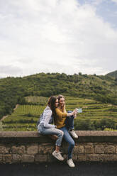 Zwei junge Frauen sitzen auf einer Mauer in einer ländlichen Landschaft und machen ein Selfie, Greve in Chianti, Toskana, Italien - JPIF00566