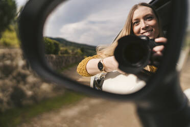 Reflection in wing mirror of smiling young woman with camera leaning out of car window - JPIF00578