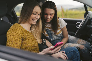 Zwei glückliche junge Frauen auf einem Roadtrip, die ihr Mobiltelefon teilen - JPIF00582
