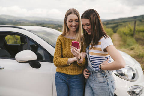 Two happy young women standing beside car sharing cell phone stock photo