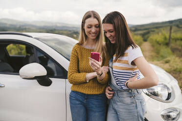 Zwei glückliche junge Frauen stehen neben einem Auto und teilen sich ein Mobiltelefon - JPIF00583