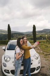 Zwei glückliche junge Frauen stehen neben einem Auto und machen ein Selfie, Toskana, Italien - JPIF00584