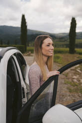 Lächelnde junge Frau beim Aussteigen aus dem Auto in ländlicher Landschaft, Toskana, Italien - JPIF00585