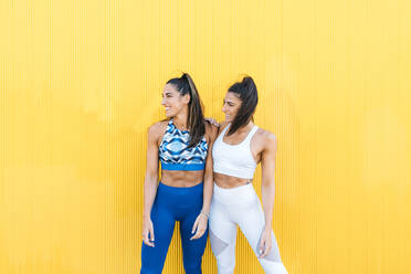 Smiling sportive twins posing in front of a yellow and pink wall - DAMF00280