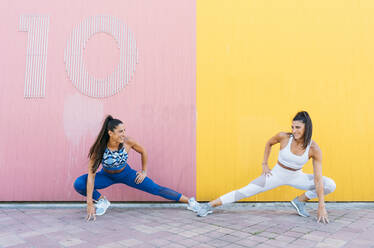 Lächelnde, sportliche Zwillinge, die ihre Beine vor einer gelb-rosa Wand ausstrecken - DAMF00281
