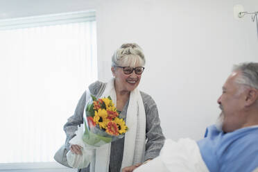 Happy senior woman bringing flower bouquet to husband recovering in hospital room - CAIF24735