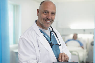 Porträt eines selbstbewussten, lächelnden männlichen Arztes, der ein digitales Tablet in einem Krankenhauszimmer benutzt - CAIF24748
