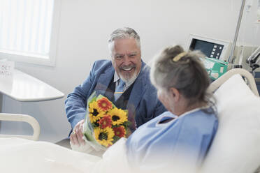 Glücklicher älterer Mann mit Blumenstrauß, der seine Frau im Krankenhaus besucht - CAIF24750