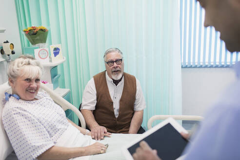 Arzt mit digitalem Tablet bei der Visite, Gespräch mit älterem Ehepaar im Krankenhauszimmer - CAIF24790
