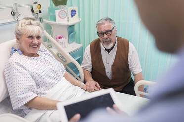 Arzt mit digitalem Tablet im Gespräch mit einem älteren Ehepaar im Krankenhauszimmer - CAIF24801