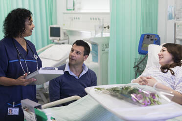 Doctor with medical chart making rounds, talking with couple in hospital room - CAIF24822