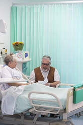 Älterer Mann mit Glückwunschkarte, der seine im Krankenhaus liegende Frau besucht - CAIF24829