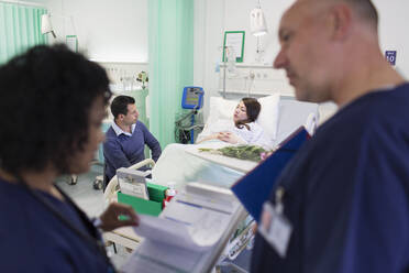 Ärzte mit Krankenblatt machen Visite, während der Patient im Krankenhausbett im Hintergrund ruht - CAIF24840
