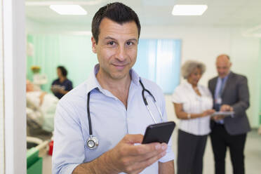 Porträt eines selbstbewussten männlichen Arztes, der ein Smartphone in einer Krankenstation benutzt - CAIF24853