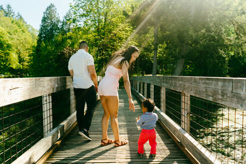 Blick von hinten auf eine Familie, die auf einer Brücke spazieren geht, mit Sonnenflecken - CAVF77635