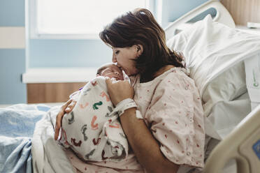 Side view of mother in hospital bed kissing newborn son's head - CAVF77670