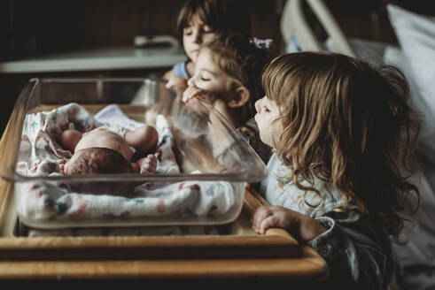 Drei Geschwister betrachten ihren neugeborenen Bruder im Stubenwagen des Krankenhauses - CAVF77720