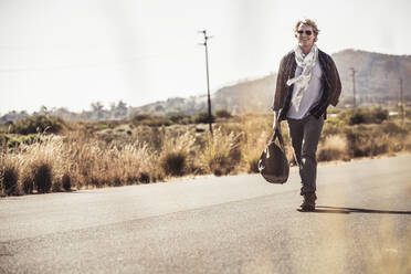 Smiling man with travelling bag walking on country road - SDAHF00669