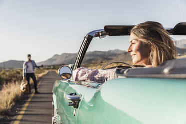 Woman in convertible car on a road trip with man hitchhiking at the roadside - SDAHF00681