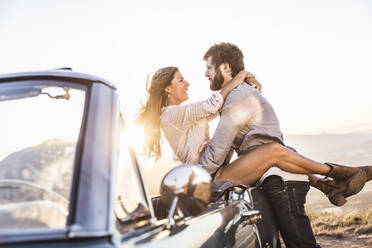 Happy affectionate couple at convertible car in the countryside at sunset - SDAHF00716