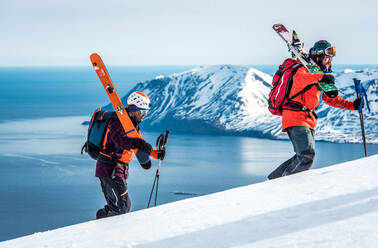 Men hiking uphill with skis and ocean and mountain behind them - CAVF77847