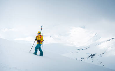 Eine Frau lächelt, während sie mit Skiern auf dem Rücken einen Hang hinaufwandert - CAVF77887