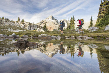 Side view and reflection of backpackers hiking beside alpine lake. - CAVF77941