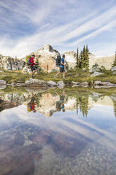 Side view and reflection of backpackers hiking beside alpine lake. - CAVF77944