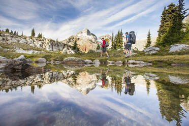 Reflection of backpackers hiking beside alpine lake in mountains. - CAVF77945