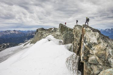 Three climbers stand on a rocky summit above snow covered glacier. - CAVF77966