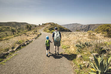 Rückenansicht von Vater und kleinem Sohn mit Rucksäcken, die Hand in Hand auf einer Landstraße gehen, La Gomera, Kanarische Inseln, Spanien - IHF00292
