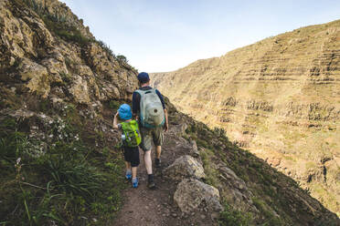 Back view of father and little son with backpacks walking on a hiking trail in the mountains, La Gomera, Canary Islands, Spain - IHF00294