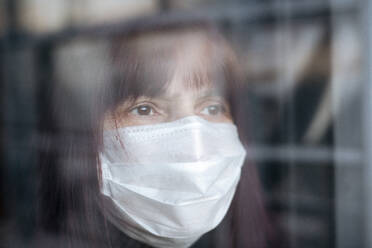 Porträt einer älteren Frau, die zu Hause eine Maske trägt und aus dem Fenster schaut - EYAF00997