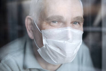 Portrait of senior man wearing a mask at home, looking out of the window - EYAF00999