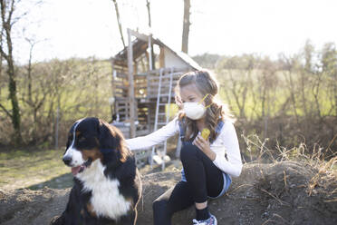 Mädchen mit Gesichtsmaske, sitzt im Garten und spielt mit ihrem Hund - HMEF00839