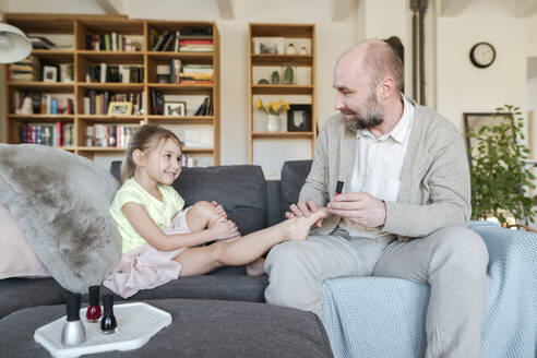 Vater lackiert die Zehennägel seiner Tochter mit rotem Nagellack auf der Couch im Wohnzimmer - VYF00089