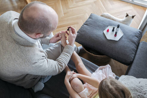 Vater lackiert die Zehennägel seiner Tochter mit rotem Nagellack auf der Couch im Wohnzimmer, Draufsicht - VYF00091