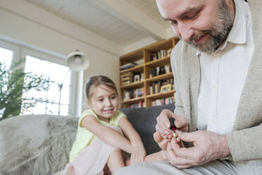 Vater lackiert die Zehennägel seiner Tochter mit rotem Nagellack auf der Couch im Wohnzimmer - VYF00093