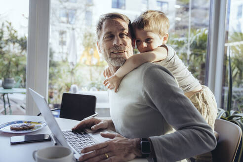 Kleiner Junge, der seinen Vater von hinten umarmt, während er von zu Hause aus arbeitet - MFF05166