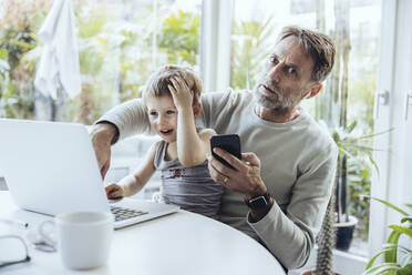 Little boy sitting on lap of father, traying to work from home - MFF05171