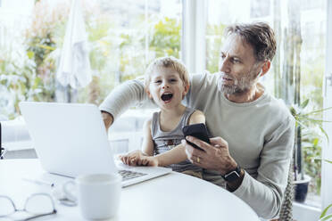 Little boy sitting on lap of father, traying to work from home - MFF05172