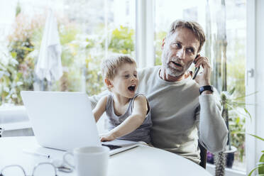 Little boy sitting on lap of father, traying to work from home - MFF05173