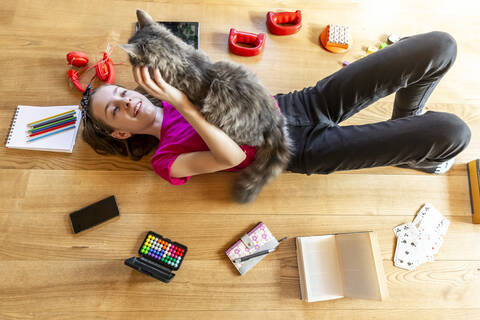 Mädchen auf dem Boden liegend, Katze kuschelnd, umgeben von Spielgeräten, lizenzfreies Stockfoto