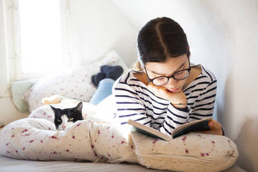 Girl lying on bed with cat reading a book - LVF08699