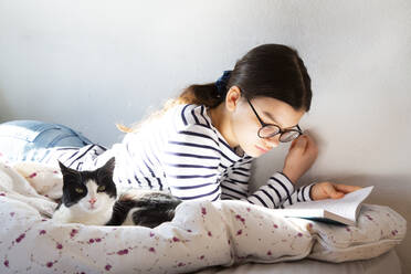 Girl lying on bed with cat reading a book - LVF08700