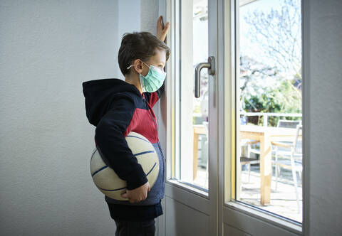 Sad boy with basketball and mask looking out of window stock photo