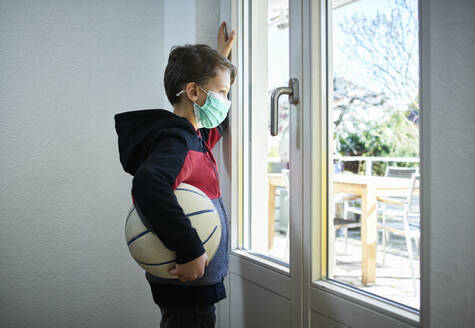 Sad boy with basketball and mask looking out of window - DIKF00405