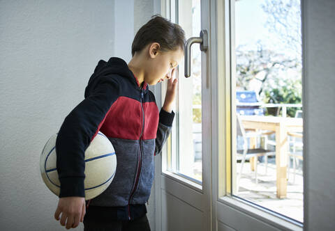 Trauriger Junge mit Basketball, der sich gegen das Fenster lehnt, lizenzfreies Stockfoto