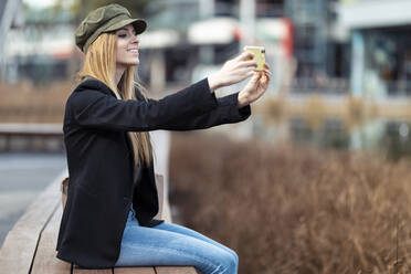 Lächelnde junge Frau sitzt auf einer Bank und macht ein Selfie mit ihrem Smartphone - JSRF00944