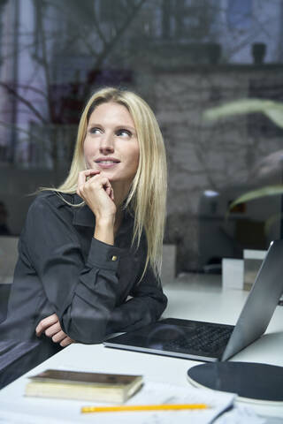 Portrait of smiling blond woman with laptop behind windowpane in office stock photo